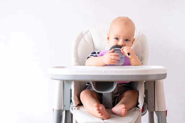 Comida para bebés. pequeño niño caucásico bebe agua de la taza de plástico verde en la trona sobre fondo blanco. Niños recién nacidos. Espacio de copia — Foto de Stock