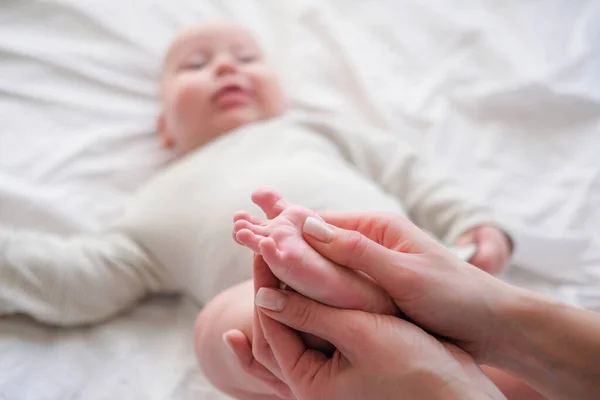 Pies de bebé en manos de madre. Mujer joven caucásica hace masaje para el bebé feliz en la cama blanca en casa. Cuidado de niños, deporte y maternidad feliz . — Foto de Stock