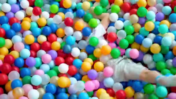 Menino loiro espalha bolas em lados diferentes, deitado em bolas plásticas multicoloridas em grande piscina de remo seco no centro de jogo. A sorrir para a câmara. A divertir-se na sala de jogos. Actividades de lazer — Vídeo de Stock
