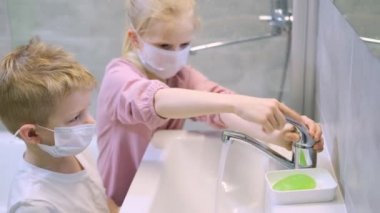 Sarışın çocuk ve kız banyoda. Tıp maskeli çocuklar modern banyoda ellerini yeşil sabunla yıkıyorlar. Temizlik, hijyen ve önleme. 4k görüntü