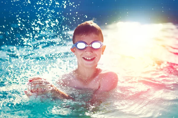 Happy little boy with goggles splashing in swimming pool