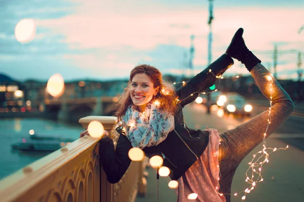 Happy young redhead woman doing yoga with fairy lights outdoors