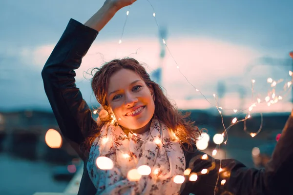 Happy young redhead woman playing with fairy lights outdoors and