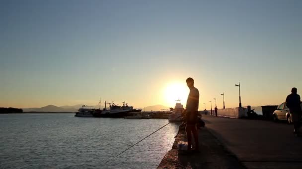 Tiro silhueta de um menino pescando no cais ao pôr do sol — Vídeo de Stock