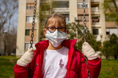 Tıbbi maskeli ve eldivenli küçük kız COVID-19 virüsünden korunmak için tek başına sallanıyor.. 