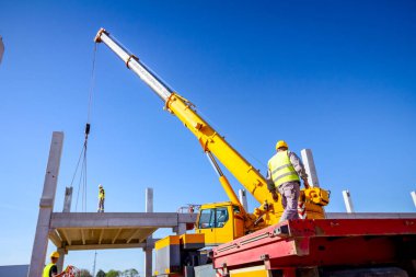 Güvenlik yeleği ve sarı miğferi olan inşaat işçisinin arkasından, inşaat için beton kirişi yöneten mobil vinç görüntüsü.