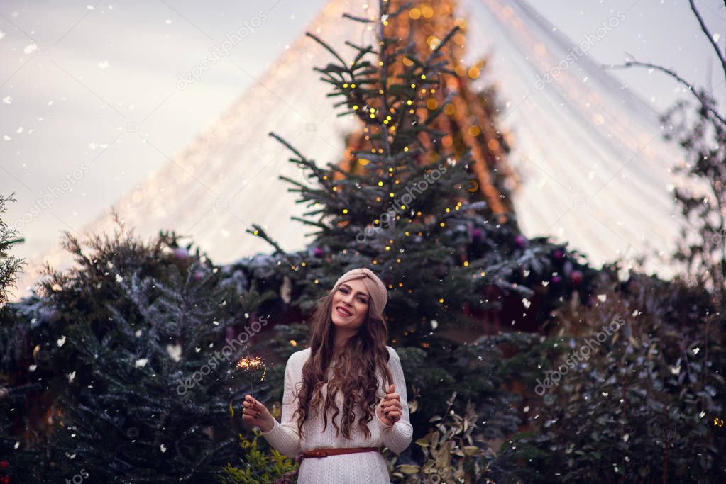 christmas portrait of happy child holding burning sparkler or firework outdoor, snowy winter decorated tree on background. New Year Holidays in city.