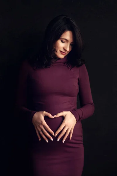Hamile mutlu kadın karnına dokunuyor. Hamile orta yaşlı anne portresi, karnını okşuyor ve yakından gülümsüyor. Sağlıklı hamilelik konsepti, siyah arka planda beklenen esmer kadın.