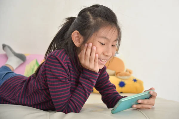 Heureuse fille asiatique enfant regardant le téléphone intelligent sur le lit à la maison . — Photo