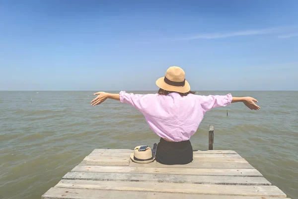 Donna che alza le mani e siede sul molo di legno di fronte al mare — Foto Stock