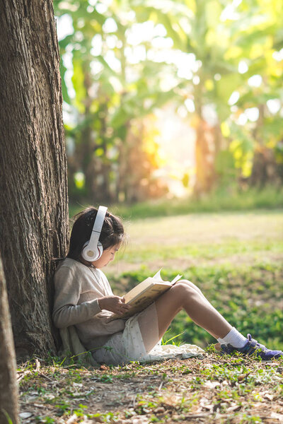 Asian girl with book and headphone siting under the big tree in 