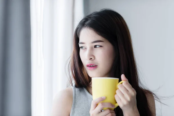 Mulher asiática segurando caneca amarela perto da janela com luz natural . — Fotografia de Stock