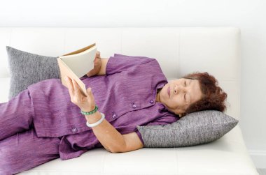 Asyalı üst düzey kadın kanepede döşenmesi ve kahverengi bir kitap okuma