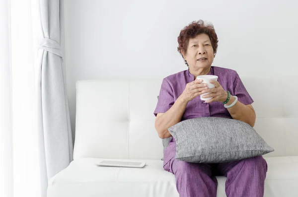 Asiática senior mujer beber agua, sentado en sofá cerca de ventana a — Foto de Stock