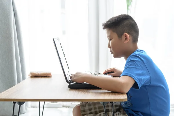 Asiático menino jogar jogo por laptop perto da janela em casa . — Fotografia de Stock