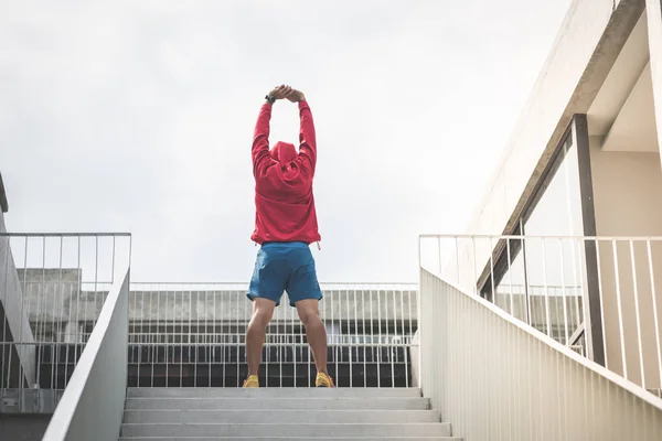 Esportista em capuz vermelho fazendo exercício perto dos degraus ao ar livre — Fotografia de Stock