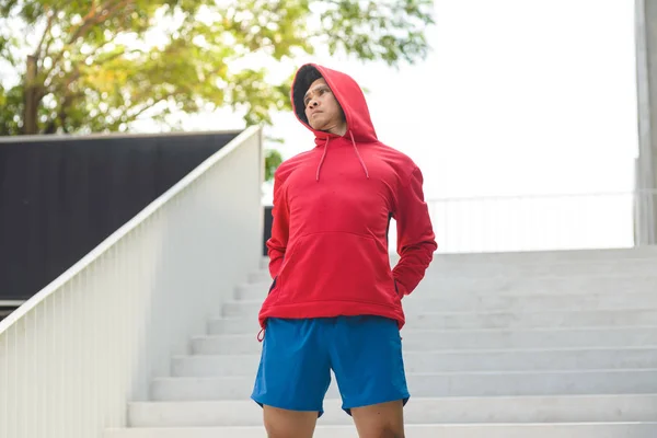 Человек спортивного телосложения в красном капюшоне согревает тело у ступенек, образ жизни — стоковое фото