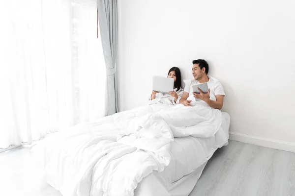 Lyckliga asiatiska paret använder laptop och tablett tillsammans på sängen på ho — Stockfoto