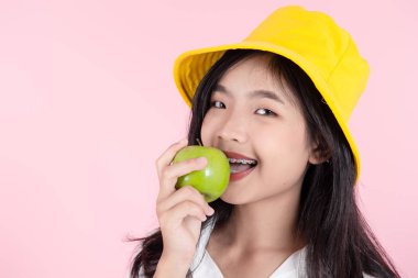 Asian teenager wearing yellow hat eats green apple on pink backg