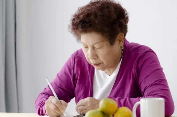 Ασιάτισσα ηλικιωμένη γυναίκα γράφει σημειωματάριο στο σπίτι, lifestyle έννοια. — Φωτογραφία Αρχείου