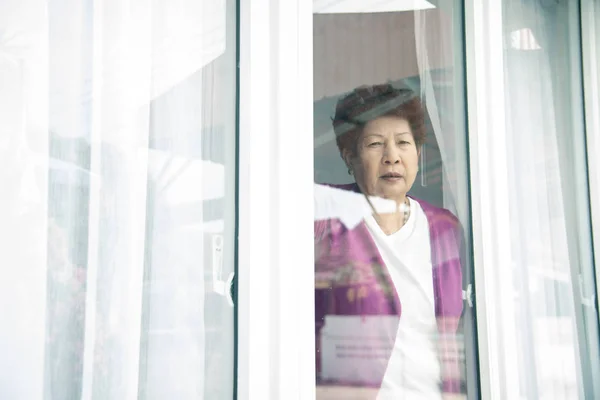 Aziatisch senior vrouw staan in de buurt van venster en op zoek outdoor. — Stockfoto