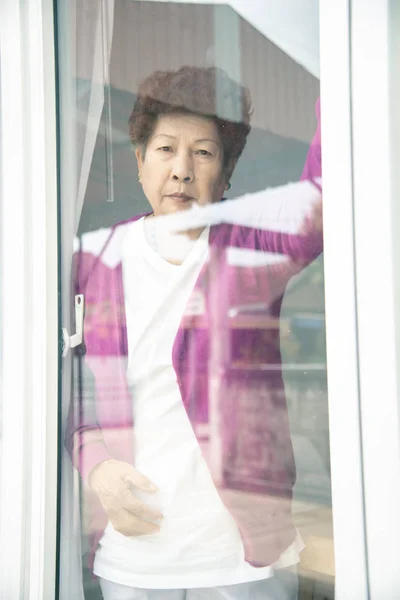 Asiática senior mujer de pie cerca de ventana y mirando al aire libre . —  Fotos de Stock