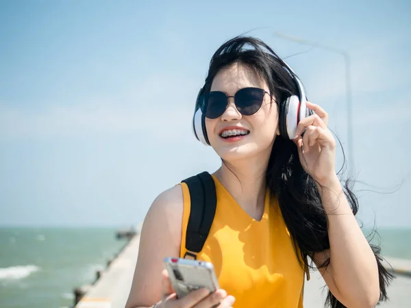 Heureuse femme asiatique avec sac à dos en utilisant casque sur la plage, li — Photo