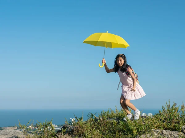 Girl with backpack holding yellow umbrella climbing on the top o — Stok fotoğraf