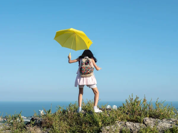 Girl with backpack holding yellow umbrella climbing on the top o — Foto Stock