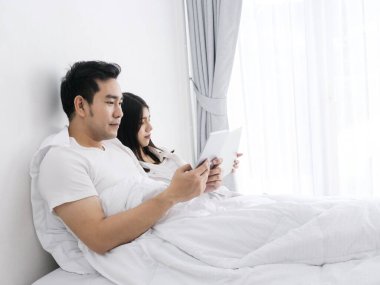 Mutlu Asyalı çift yatakta tablet ve dizüstü bilgisayar kullanıyor, evde çalışıyor, evde kalıp güvende kalıyorlar..