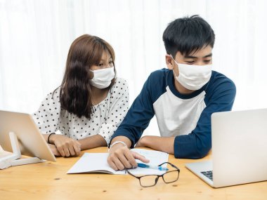 Maske takan Asyalı bir genç evden online eğitim için dizüstü bilgisayar ve tablet kullanıyor, karantina konsepti.