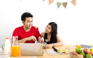 Evde dizüstü bilgisayar kullanan, salata yiyen ve taze meyve yiyen mutlu bir Asyalı çift. Evde kal. Dikkatli ol..