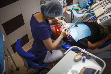 Tulumlu dişçi, yüz maskesi, kep ve lastik eldiven. Kadın dişçi, dişçi koltuğundaki hastayı tedavi eder..