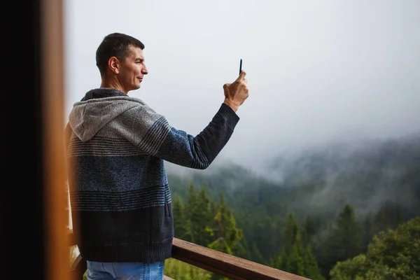 Genç bir adam telefonuyla otelin balkonundan dağlara bakan güzel bir manzaranın fotoğraflarını çekiyor. Bulgaristan 'ın Pamporovo kayak merkezindeki otelde köknar ağaçları, sis ve dağların güzel manzarası