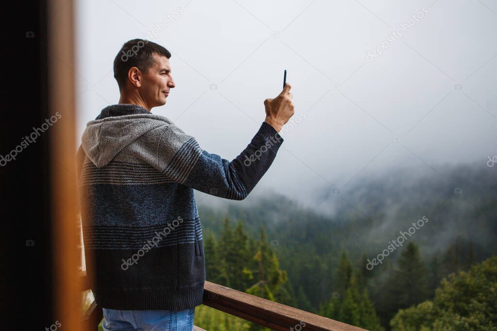 A young man takes photos with his phone of a beautiful view from the hotel's balcony overlooking the mountains. Beautiful view from the window of fir trees, fog and mountains in the hotel in the Pamporovo ski resort in Bulgaria