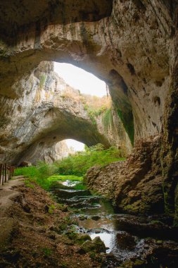Tepesinde yuvarlak delikler olan büyük taş bir mağaranın yüksek kemerleri, mağaranın içinde çitleri olan turistik bir yol. Bulgaristan 'ın doğal cazibe-Devetashka mağarası