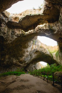 Tepesinde yuvarlak delikler olan büyük taş bir mağaranın yüksek kemerleri, mağaranın içinde çitleri olan turistik bir yol. Bulgaristan 'ın doğal cazibe-Devetashka mağarası