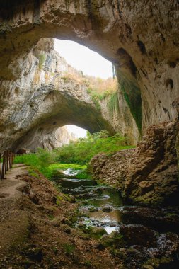 Tepesinde yuvarlak delikler olan büyük taş bir mağaranın yüksek kemerleri, mağaranın içinde çitleri olan turistik bir yol. Bulgaristan 'ın doğal cazibe-Devetashka mağarası