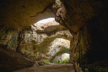 Tepesinde yuvarlak delikler olan büyük taş bir mağaranın yüksek kemerleri, mağaranın içinde çitleri olan turistik bir yol. Bulgaristan 'ın doğal cazibe-Devetashka mağarası