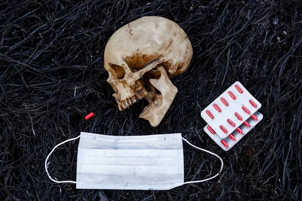 医学用医薬品の赤いカプセルと保護医療用マスクを持つ焼け草の黒い背景に人間の頭蓋骨 ワクチンの必要性や世界の危険性を象徴しています — ストック写真