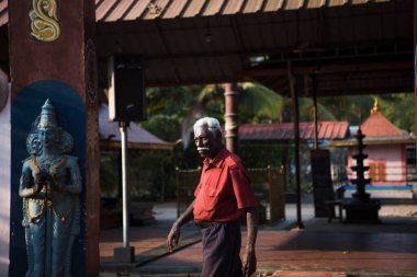 Kerala, Hindistan-3 Şubat 2019. Hintli yaşlı, gri saçlı, kırmızı tişörtlü bir adam yol boyunca Hindu tapınağının arka planına doğru yürüyor.