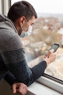 Evinin penceresinde tıbbi maske takan genç bir adam akıllı telefona bakıyor ve panoramanın fotoğraflarını çekiyor. Koyu renk saçlı bir adam, kendini izole ettiği sırada boş bir şehre koronavirüsle bakar.