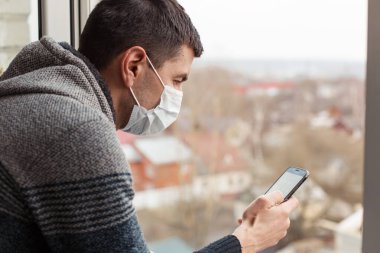 Evinin penceresinde tıbbi maske takan genç bir adam akıllı telefona bakıyor ve panoramanın fotoğraflarını çekiyor. Koyu renk saçlı bir adam, kendini izole ettiği sırada boş bir şehre koronavirüsle bakar.