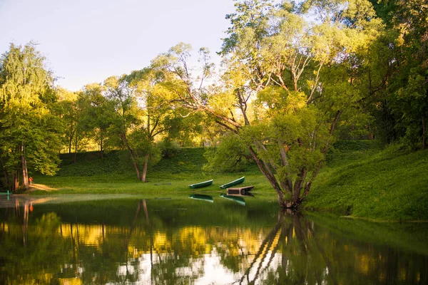 Lake in the Park among green trees and fields of dandelions. Trees are reflected in the lake and two wooden boats are standing on the shore