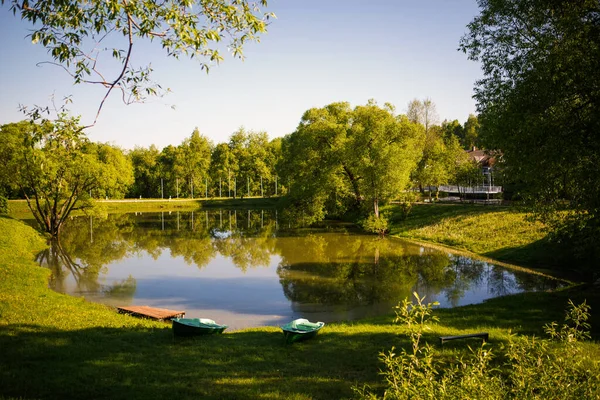 Lake in the Park among green trees and fields of dandelions. Trees are reflected in the lake and two wooden boats are standing on the shore