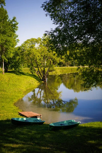 Lake in the Park among green trees and fields of dandelions. Trees are reflected in the lake and two wooden boats are standing on the shore