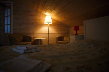 Sarı bir abajur tavan arası yatak odasında düşük tavanlı loş ve rahat bir şekilde parlar. Lamba yatağın yanında. Bir kır evinde, ahşap bir yatak odasının içinde..