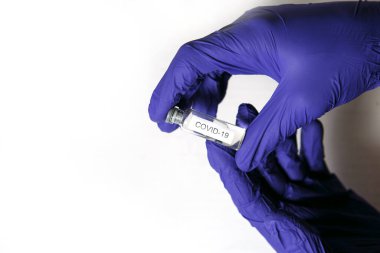 Doktorun elleri mavi tıbbi lastik eldivenlerde üzerinde COVID-19 yazan bir şişe tutuyor. Tıbbi konsept, bir şişe koronavirüs, tedavi ve aşı, salgınla mücadele. Beyaz arkaplan