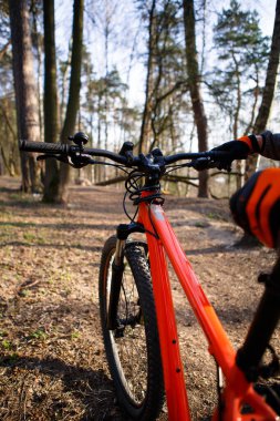 Otoyoldaki ormanda turuncu çerçeveli bisiklet. Siyah ve turuncu eldivenli bir bisikletçi motoru çerçeve ve koltuktan tutar. Bisiklet, spor, sağlıklı yaşam tarzı teması