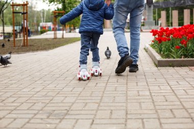Babası küçük kızına dört tekerlekli paten sürmeyi öğretiyor. Baba, çocuğun elinden, ayaklarından ve patenli çocuğundan destek alır. Yuvarlayıcılara odaklan. Babalar günü, aile hafta sonu, mesajlaşma yeri.